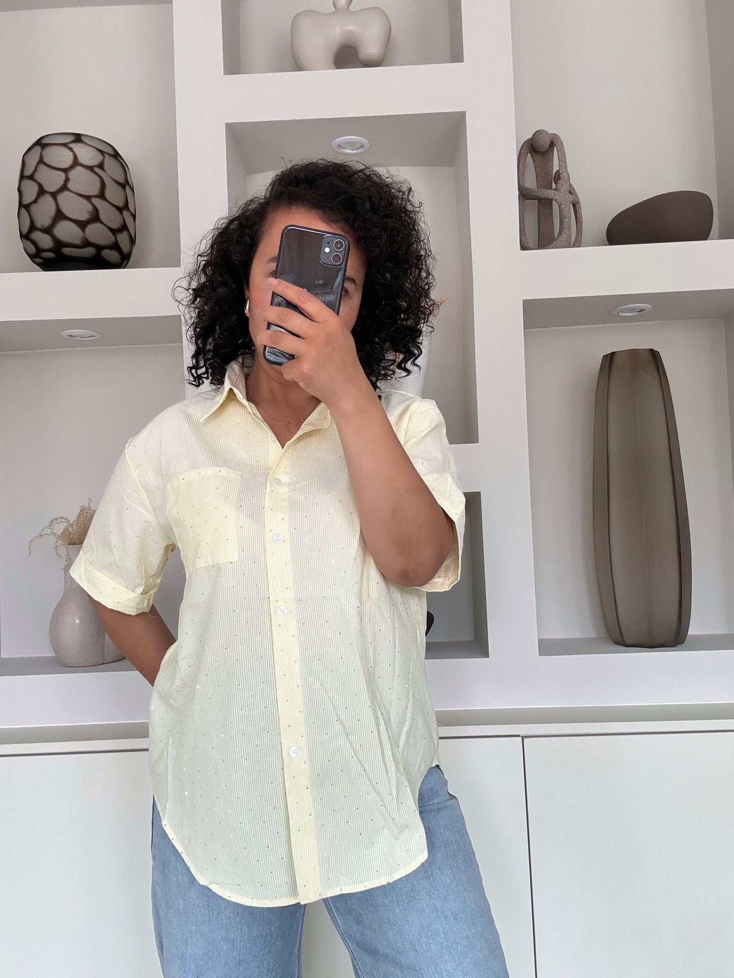 Vrouw met krullend haar draagt een lichtgele blouse met glans over een blauwe jeans, staand voor een witte kastwand met decoratie
