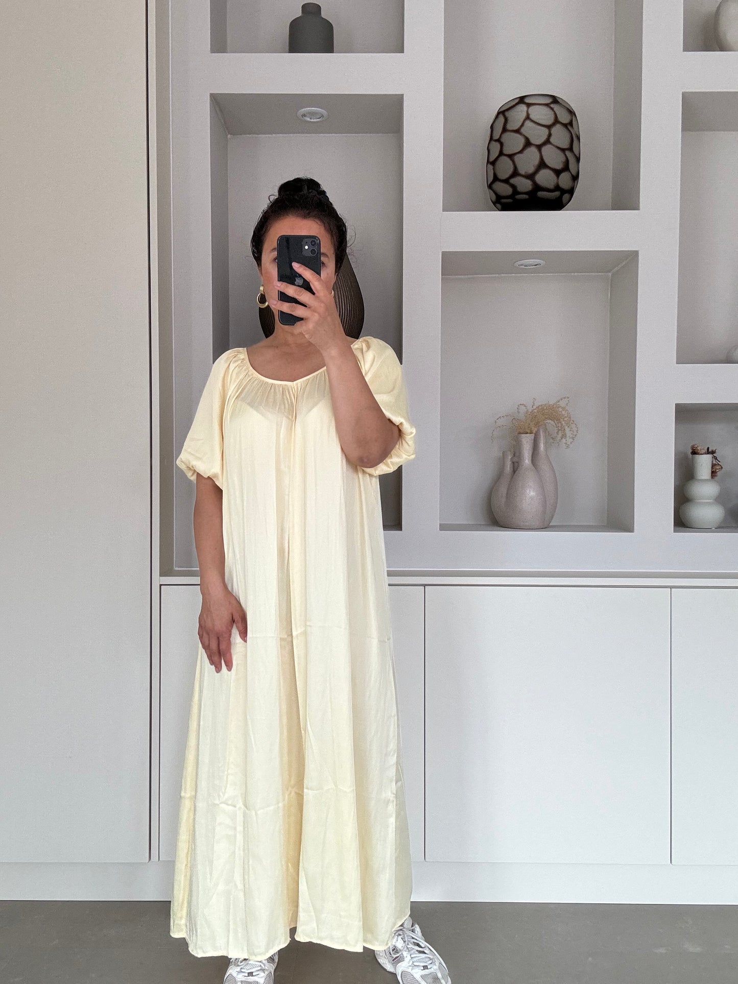 Model draagt een satijnachtige maxi-jurk in zachtgeel met korte mouwen en vloeiende pasvorm voor een elegante, luchtige look.