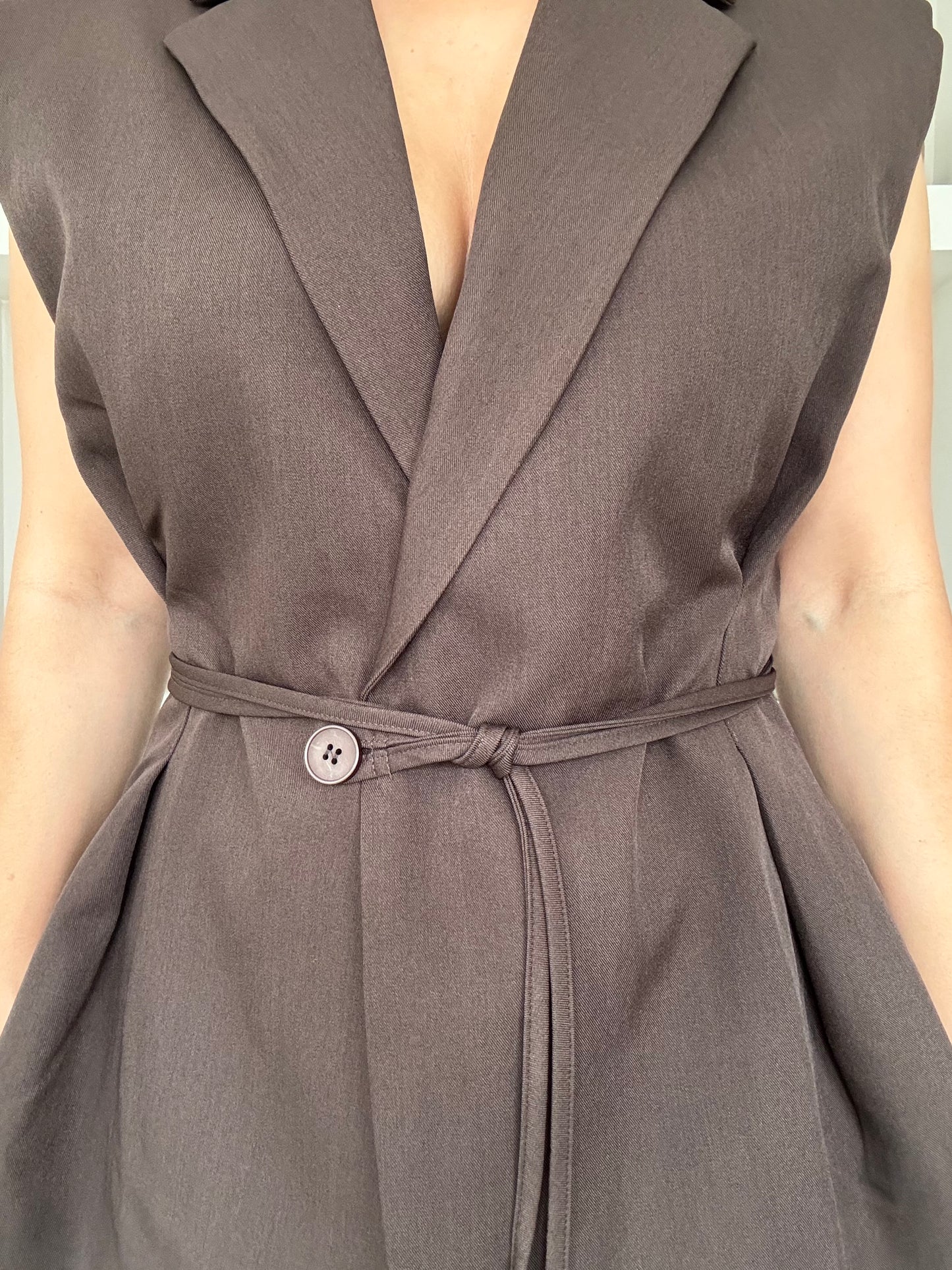 Model draagt een elegante mouwloze blazer in donkerbruin met een ceintuur in de taille, gestyled met een lichte jeans voor een minimalistische, moderne look