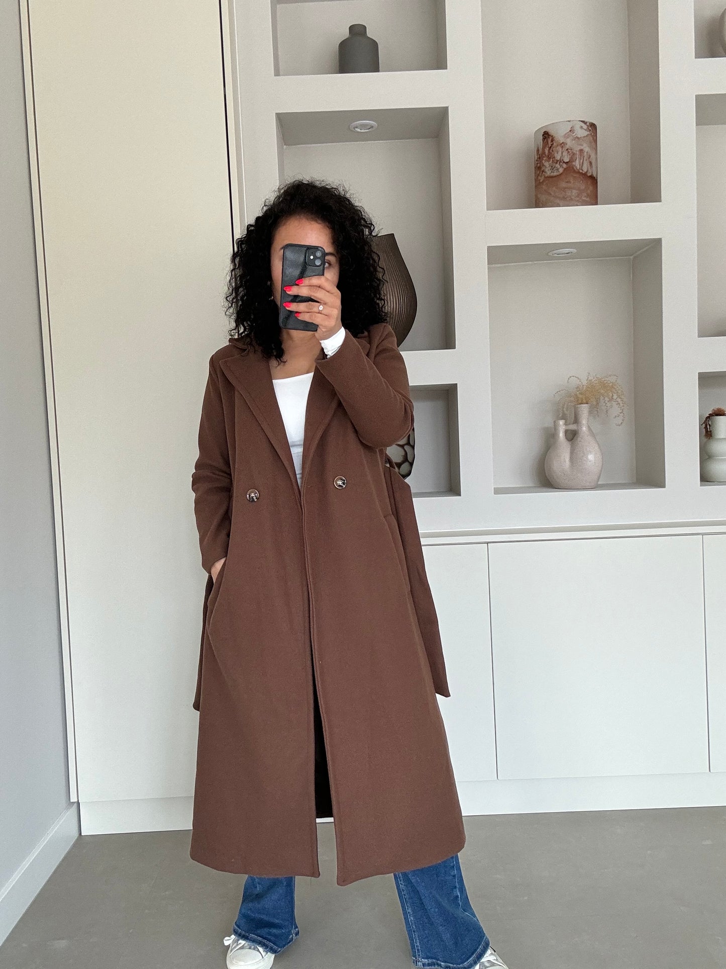 Vrouw draagt lange bruine elegante jas met dubbele knopen en diepe zakken, gecombineerd met jeans en sneakers