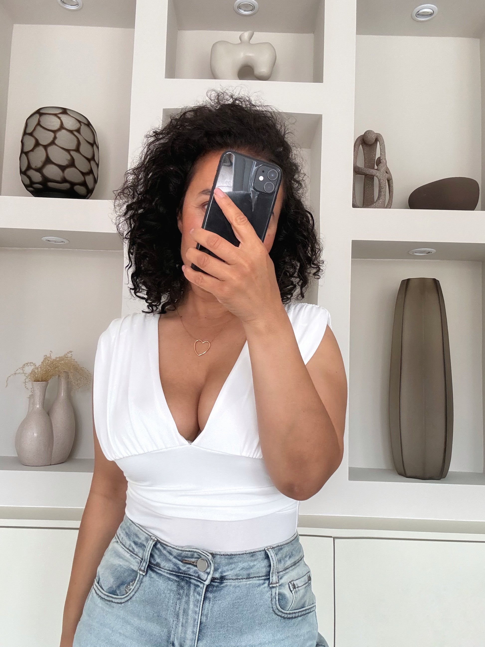 Vrouw draagt een witte V-hals top met jeans, staat voor een witte minimalistische kastwand met decoratie