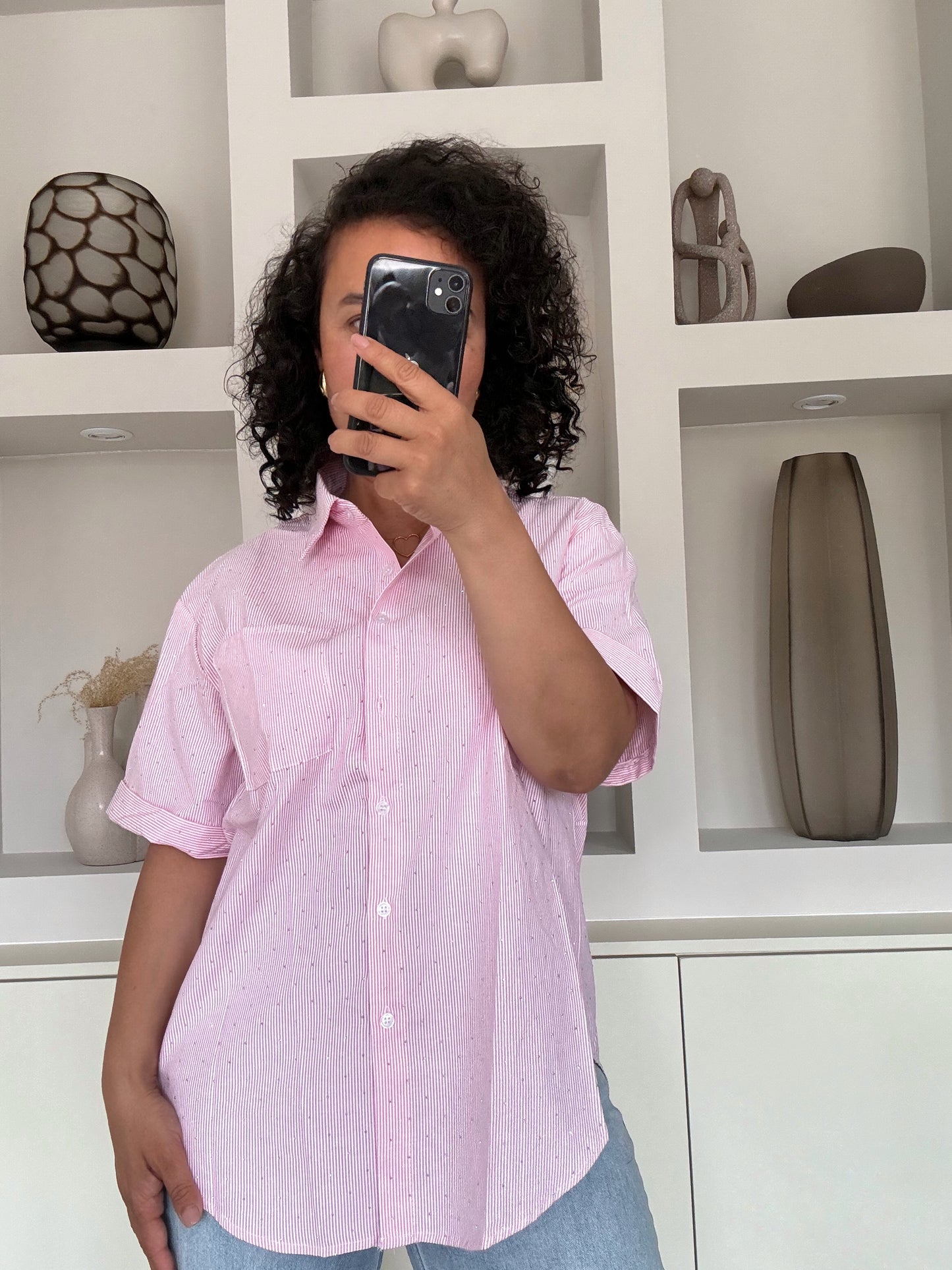 Vrouw met krullend haar draagt een lichtroze blouse met glans over een blauwe jeans, staand voor een witte kastwand met decoratie