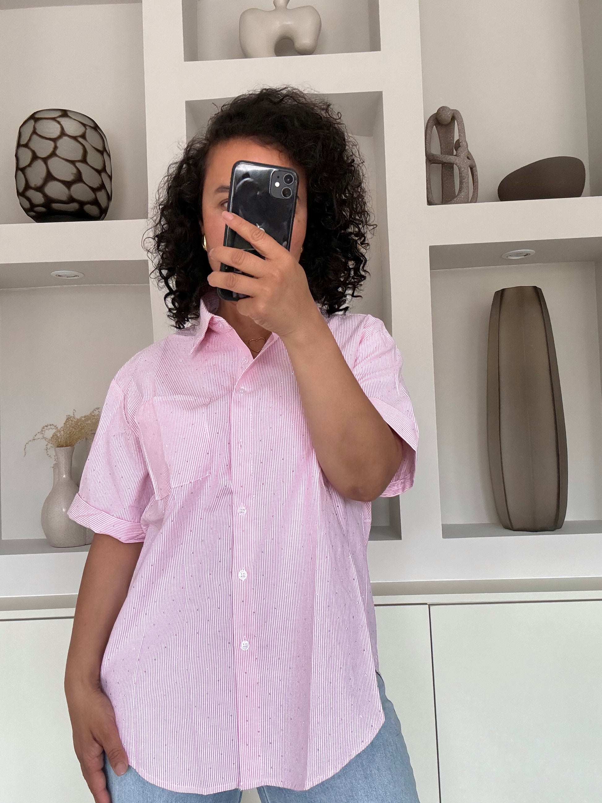 Vrouw met krullend haar draagt een lichtroze blouse met glans over een blauwe jeans, staand voor een witte kastwand met decoratie