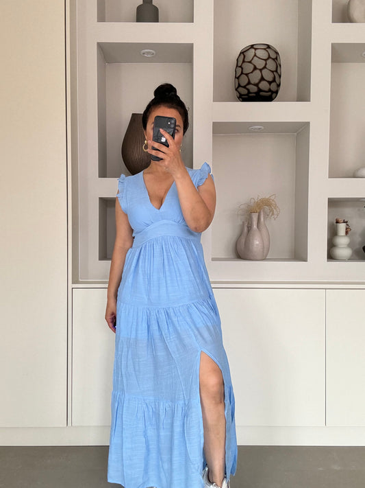 Model draagt een luchtige blauwe maxi-jurk met ruffle schouderbandjes, diepe V-hals en hoge split — perfect voor zomerse dagen.