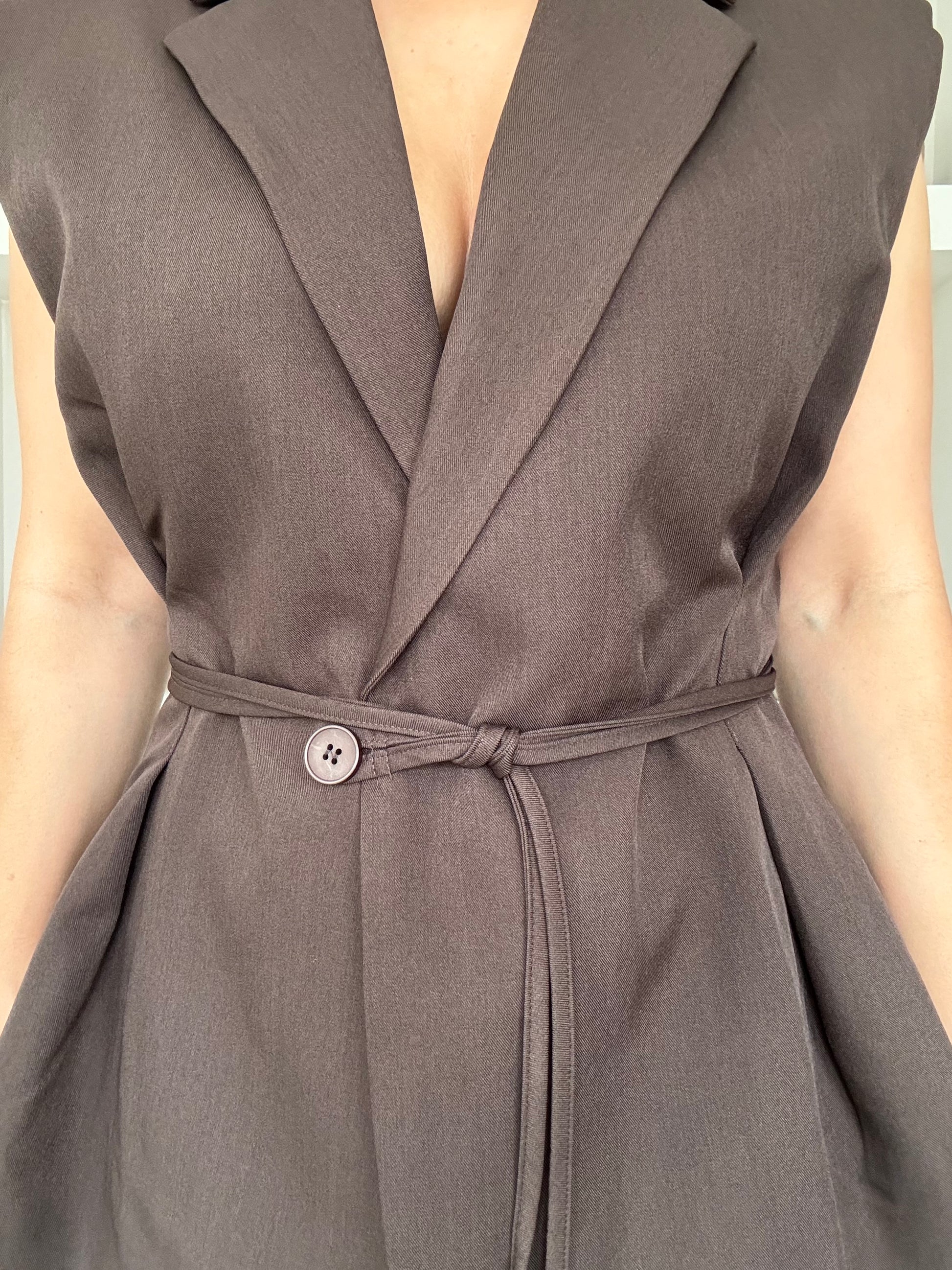Model draagt een elegante mouwloze blazer in donkerbruin met een ceintuur in de taille, gestyled met een lichte jeans voor een minimalistische, moderne look
