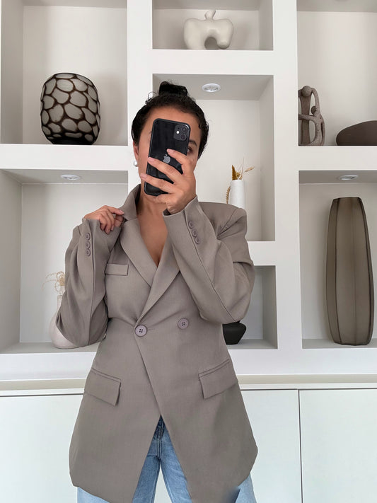 Model draagt een taupekleurige blazer met een dubbele knoopsluiting, gestructureerde schouders en een getailleerde pasvorm, gestyled met een lichte jeans