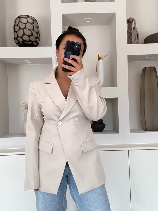Model draagt een beige blazer met dubbele knoopsluiting, gestructureerde schouders en een vrouwelijke, getailleerde pasvorm, gecombineerd met een lichte jeans.
