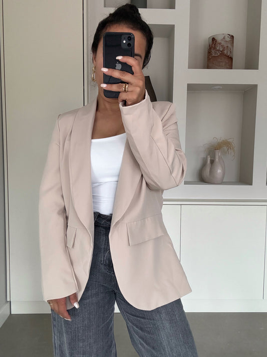Vrouw met soft-beige oversized blazer over witte top en grijze jeans in minimalistisch interieur – VYNAH