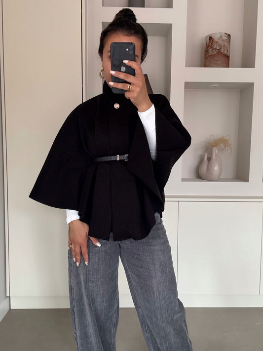 Zwarte belted cape met hoge kraag en knoopsluiting, gedragen over witte top en grijze jeans – VYNAH