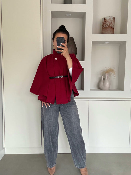 Vrouw met bordeauxrode cape met riem en hoge kraag, over witte top en grijze jeans – VYNAH
