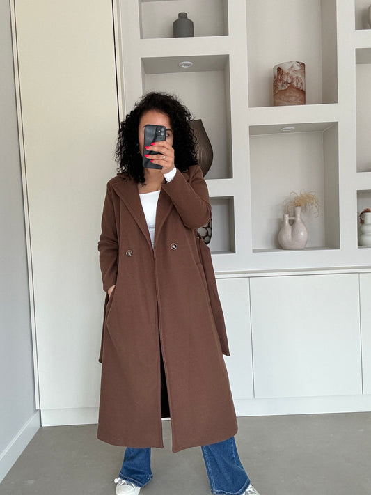 Vrouw draagt lange bruine elegante jas met dubbele knopen en diepe zakken, gecombineerd met jeans en sneakers